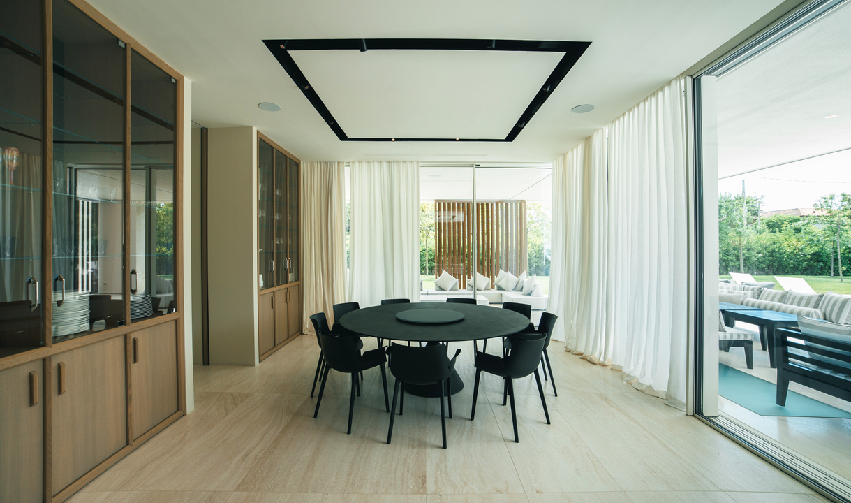 Modern dining room with a black round table, dark chairs, and a rectangular ceiling light fixture framing a recessed luminous panel.