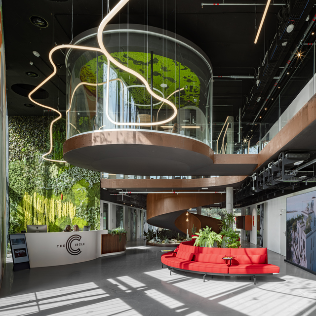 Lobby dell'edificio The Circle con lampade a sospensione LED sinuose Artemide, installazione cilindrica con parete verde, divano rosso curvo e rigogliosi giardini verticali