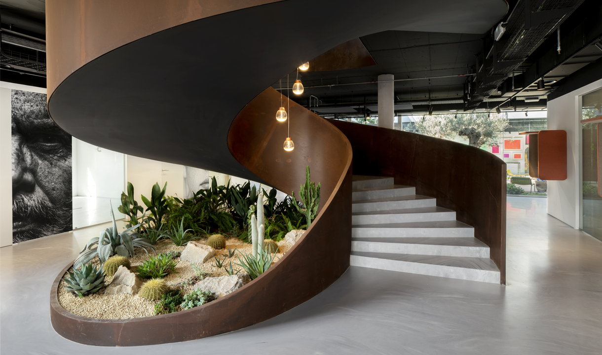 Scala a spirale con base in acciaio Corten che racchiude un giardino desertico, illuminata da tre lampadine a sospensione ambrate dall'alto.