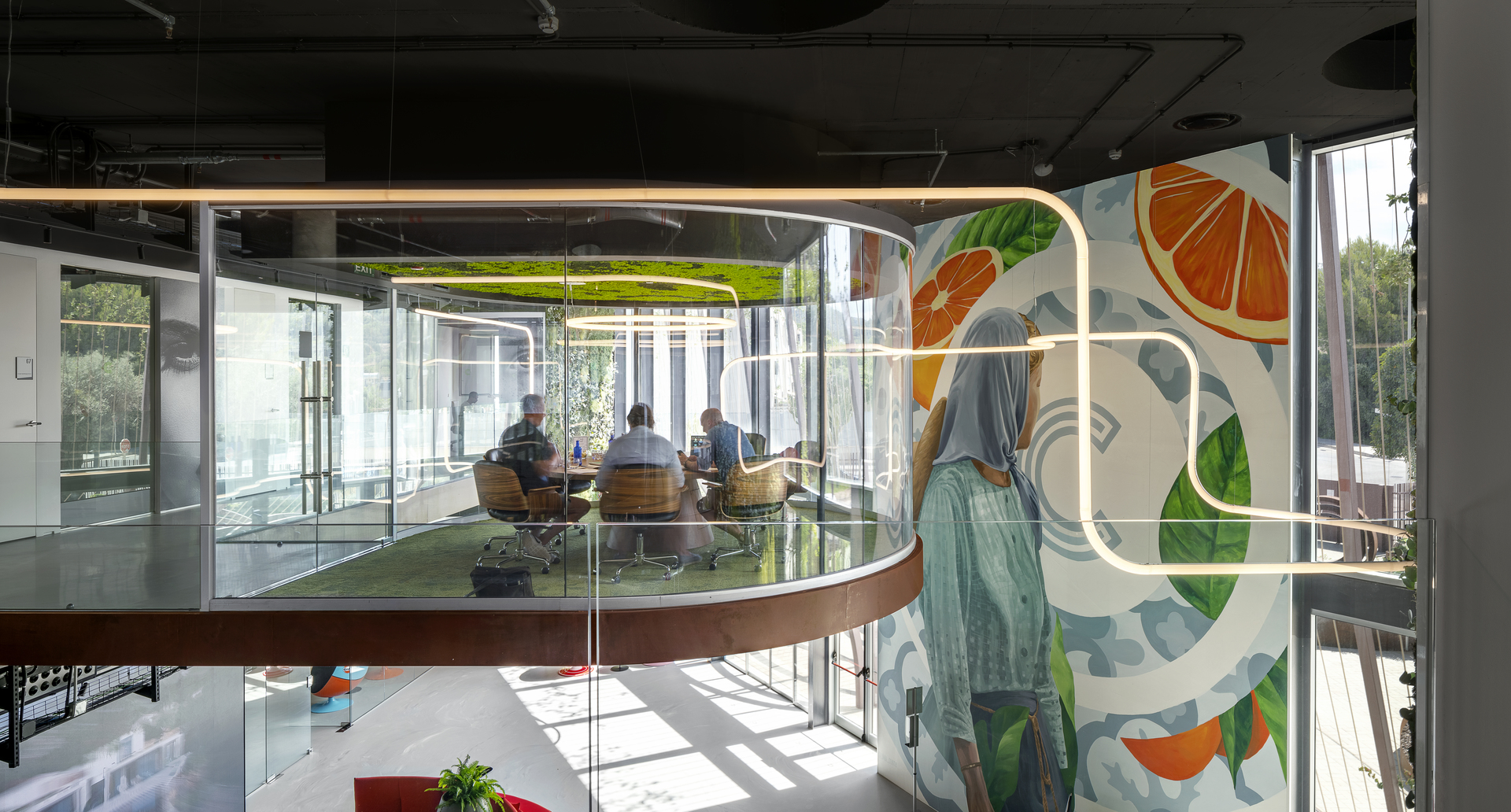 Sala riunioni moderna con pareti in vetro curve, vivace murale con frutta e illuminazione lineare stile neon Artemide sopra.