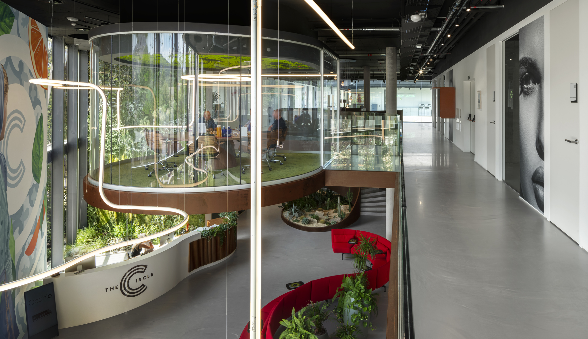 Interno di ufficio moderno con pareti in vetro curve, design biofilico, sedute rosse e lampade a sospensione lineari Artemide che illuminano lo spazio.