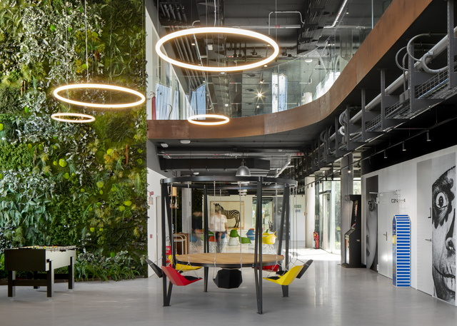 Atrio di ufficio moderno con lampade a sospensione ad anello Artemide Halo sopra un'area relax accanto a una rigogliosa parete verde.