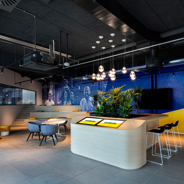 Lounge ufficio moderno con lampade a sospensione Artemide sopra una reception curva, piante, murale blu e accenti gialli.