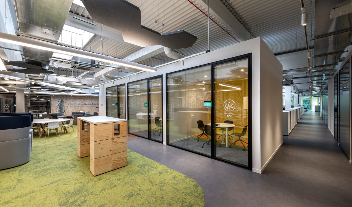Ufficio open space moderno con cabina riunioni in vetro, moquette verde, mobili contenitori in legno e soffitto industriale con illuminazione a sospensione.