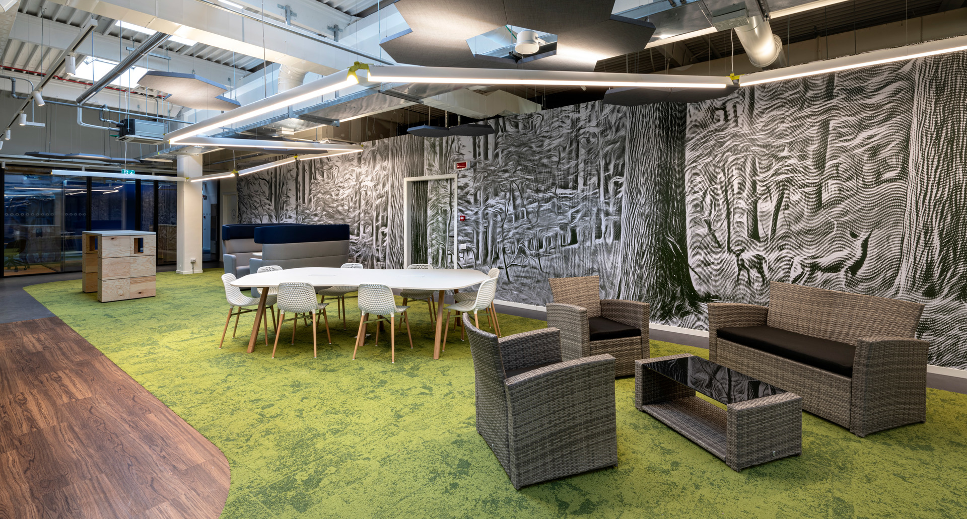 Lounge ufficio moderno con luci lineari a soffitto Artemide, sedute in vimini, moquette verde muschio e murale a tema forestale.