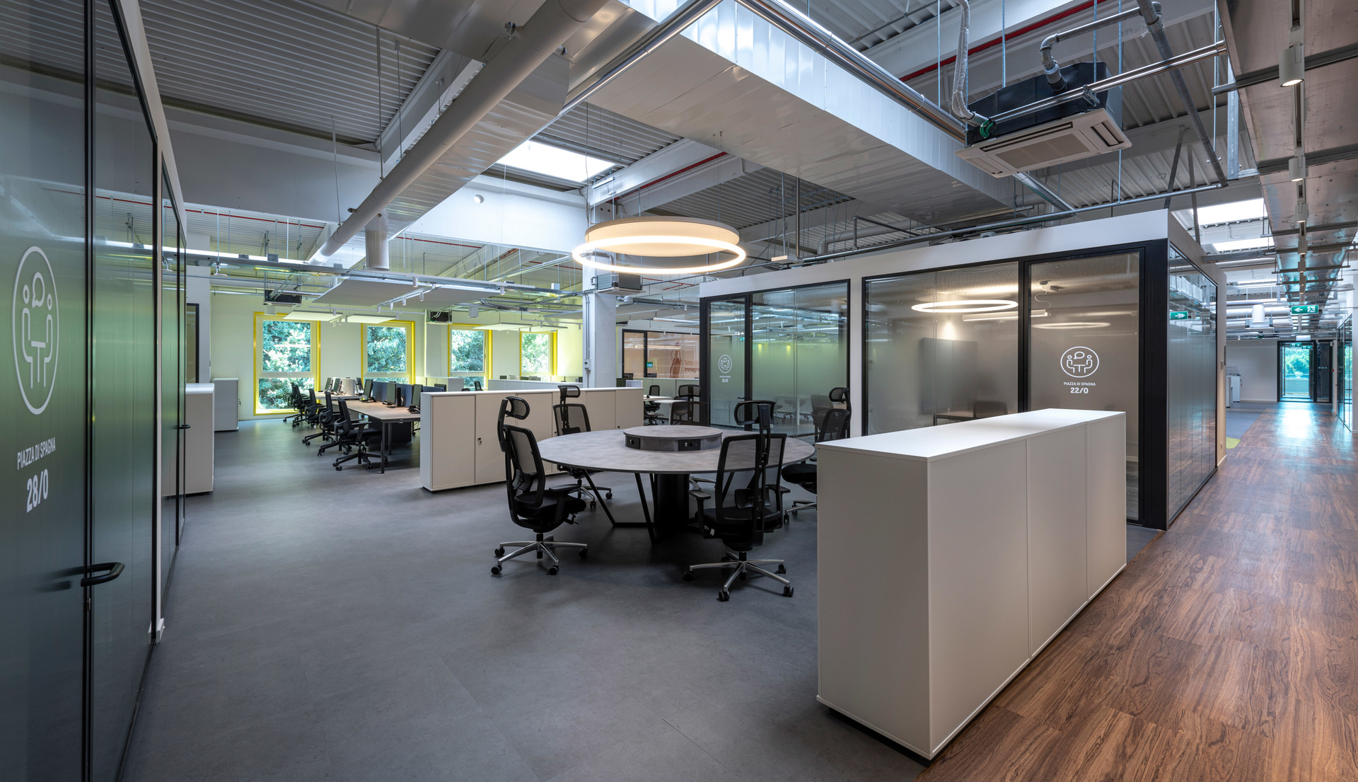 Ufficio open space moderno con lampada a sospensione circolare Artemide, sale riunioni con pareti in vetro, postazioni di lavoro e soffitto a vista in stile industriale.