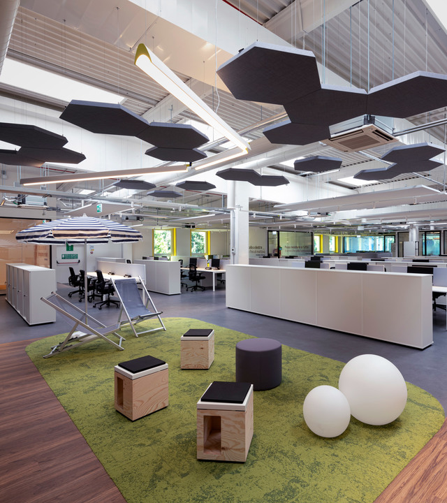 Ufficio open space moderno con pannelli acustici esagonali a soffitto, luci a sospensione bianche, area tappeto verde con sgabelli in legno e lampade da terra sferiche