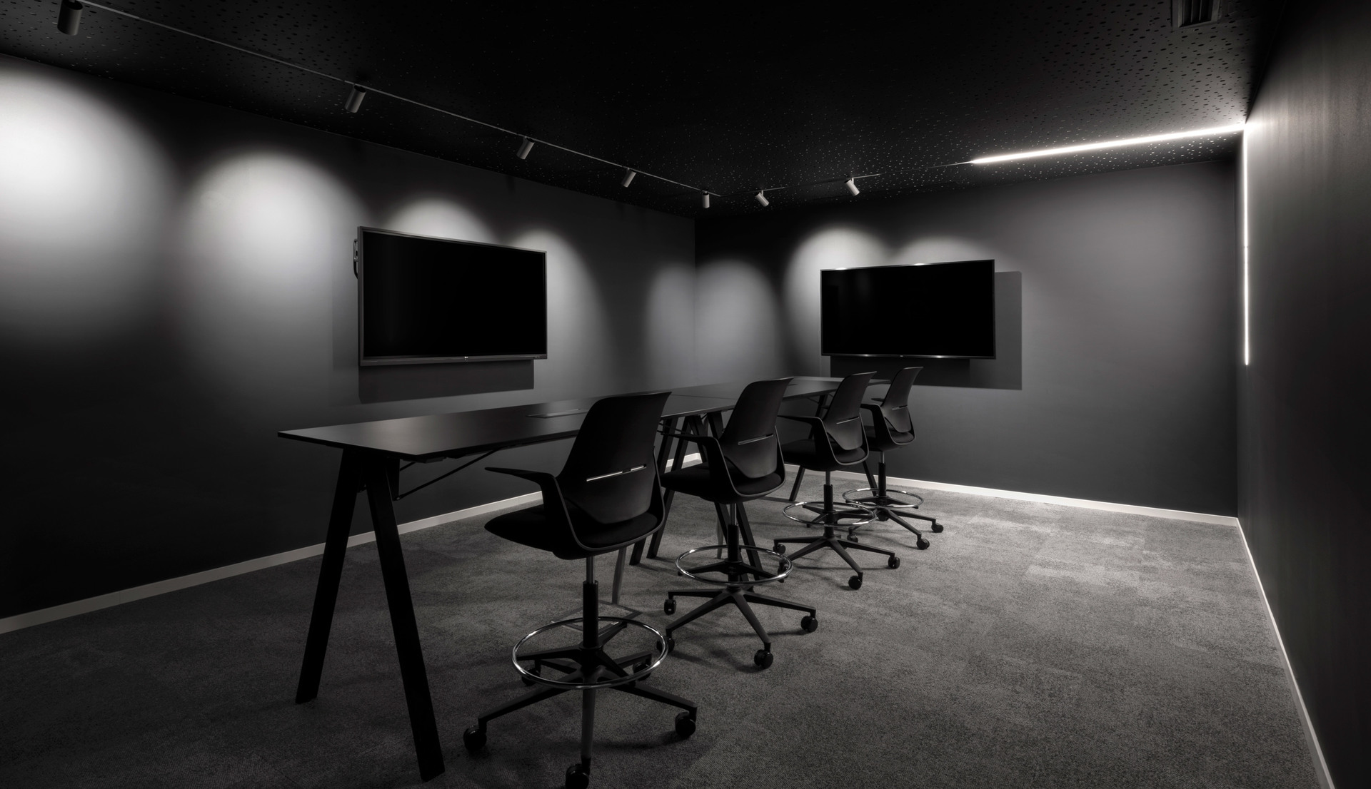 Sala riunioni scura con faretti su binario, striscia LED lineare a parete, lungo tavolo nero, sedie ergonomiche e due schermi a parete.