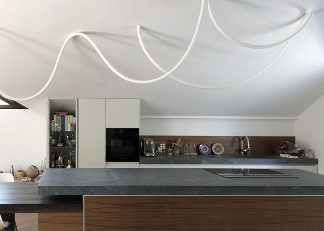 Artemide sinuous white LED tube light installation on kitchen ceiling, creating flowing curves above a modern walnut and stone island