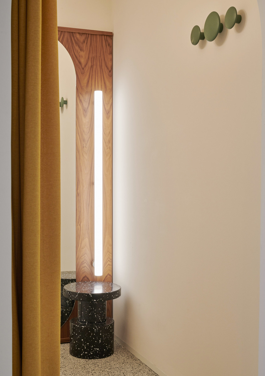 Lampada da terra verticale sottile con tubo bianco luminoso accanto a tavolino in terrazzo scuro, tenda senape calda e ganci da parete verde oliva.