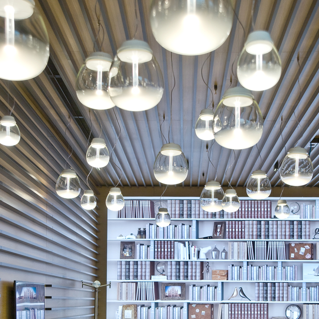 Più lampade a sospensione Artemide Empatia con globi in vetro trasparente e diffusori bianchi pendono da un soffitto a listelli sopra una libreria.