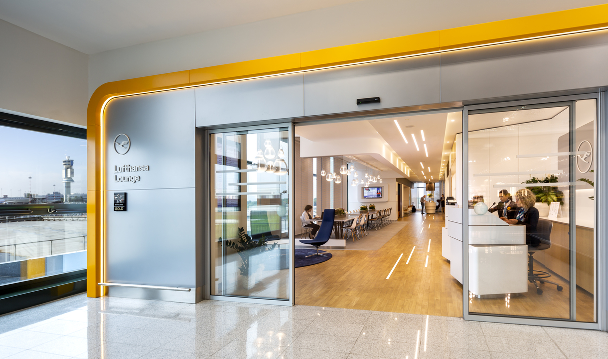 Ingresso di una lounge aeroportuale moderna con porte in vetro, accenti gialli e luci da incasso Artemide che illuminano un'area salotto.
