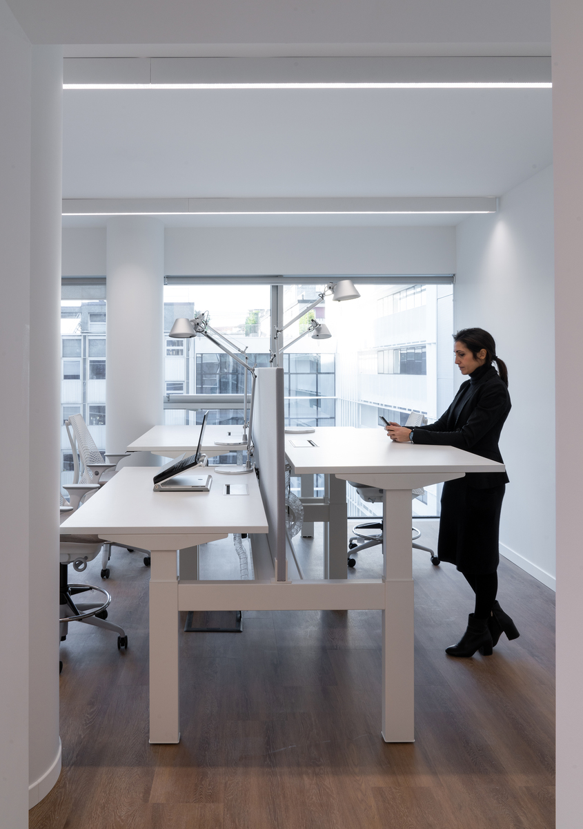 Spazio di lavoro moderno con lampade da tavolo Artemide Tolomeo su scrivanie bianche regolabili, luce lineare a soffitto e una donna che lavora in piedi.