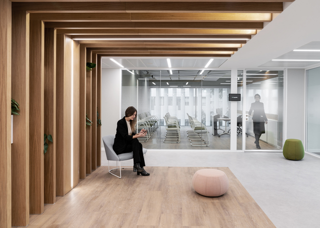 Lounge di ufficio moderno con corridoio in legno caldo, luci lineari a soffitto, pareti in vetro e una donna seduta con un tablet.