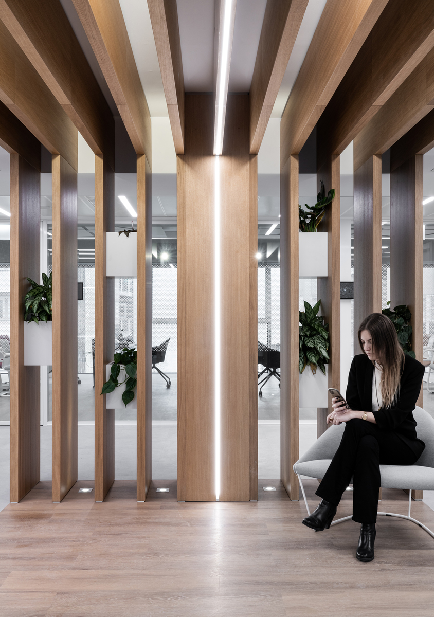 Lounge di ufficio moderno con colonne in legno con illuminazione LED lineare integrata, una donna seduta con il telefono e piante verdi.