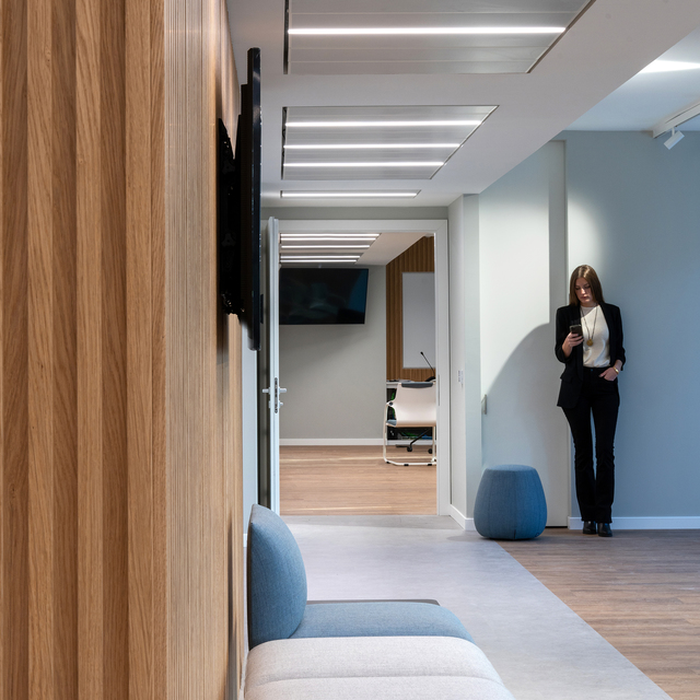 Corridoio ufficio moderno con luci LED lineari a incasso nel soffitto, parete con pannelli in legno, area salotto e donna che guarda il telefono.