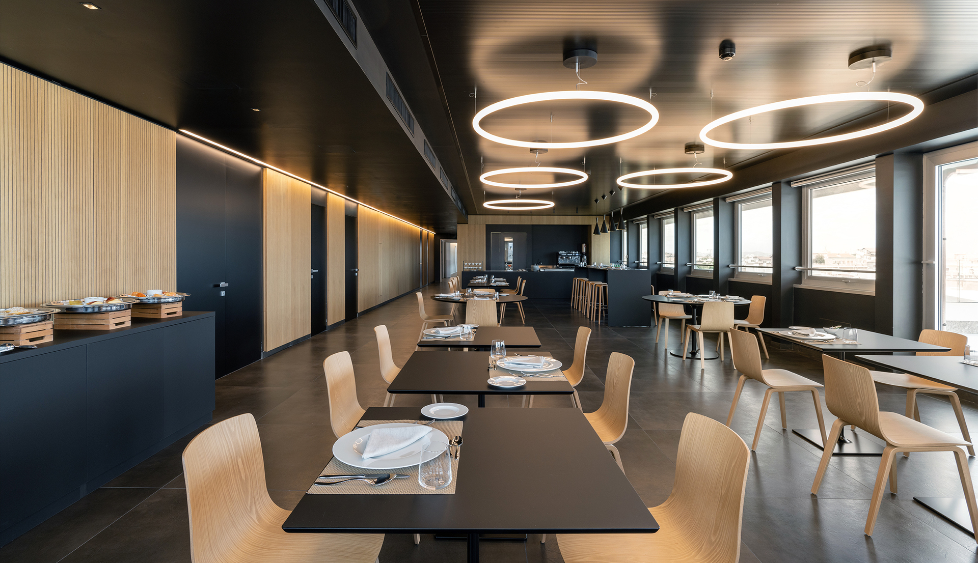 Modern restaurant with circular LED pendant lights illuminating dark ceiling above light wood chairs and black tables.