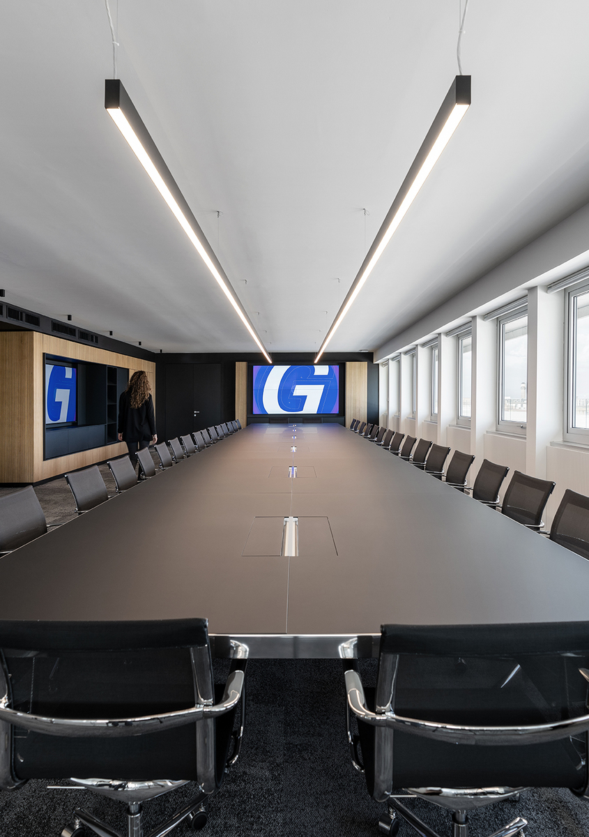 Modern conference room with a long dark table, mesh chairs, and two Artemide linear pendant lights forming a V-shape above.