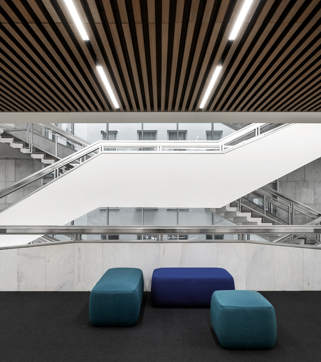 Modern atrium with linear LED lights integrated in wooden slat ceiling, marble walls, dual staircases, and colorful ottomans below.