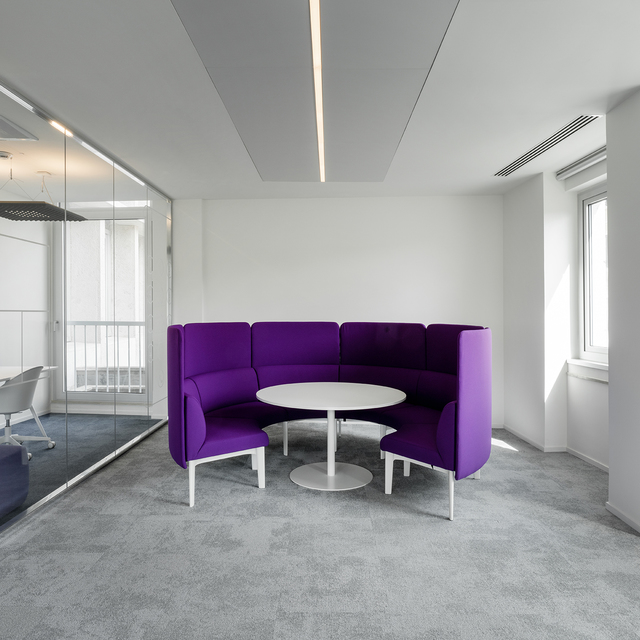 Modern office lounge with purple curved sofa and white round table, lit by Artemide recessed linear ceiling lights.
