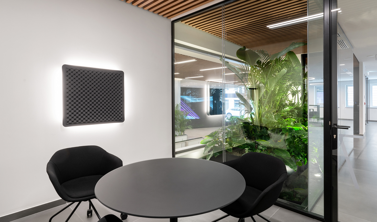 Modern office meeting area with black round table, chairs, acoustic panel with glowing edge lighting on white wall, and glass partition with plants.