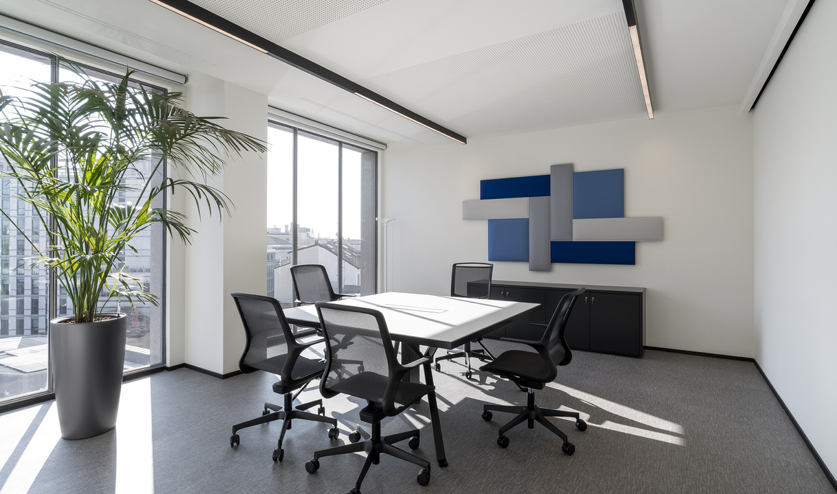 Sala riunioni moderna con luci lineari a soffitto incassate, tavolo bianco, sedie in rete nera, pannelli acustici e grande pianta da interno.