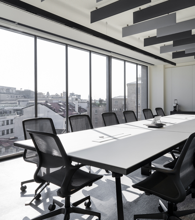 Sala conferenze moderna con lungo tavolo bianco, sedie nere a rete, finestre dal pavimento al soffitto e pannelli acustici scuri sul soffitto.