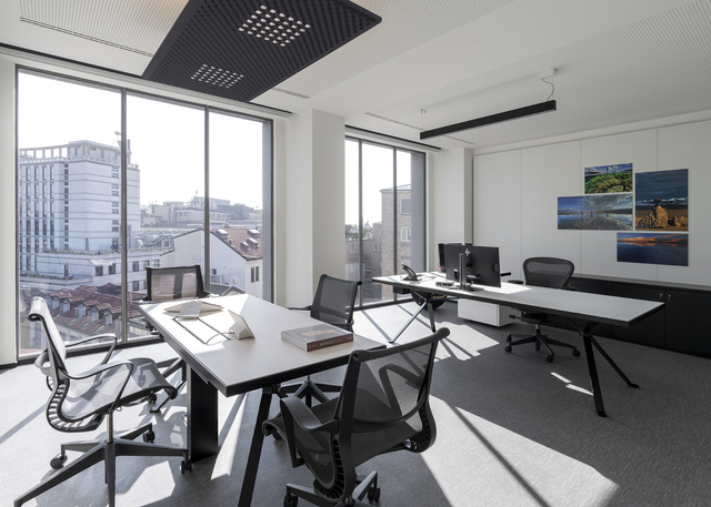 Ufficio moderno con scrivanie bianche, sedie a rete, ampie finestre e un pannello soffitto nero forato per illuminazione diffusa.