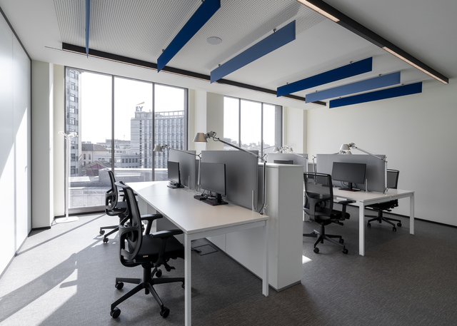 Ufficio open space moderno con scrivanie bianche, sedie operative, monitor desktop, lampade da tavolo, grandi finestre e pannelli acustici blu a soffitto.