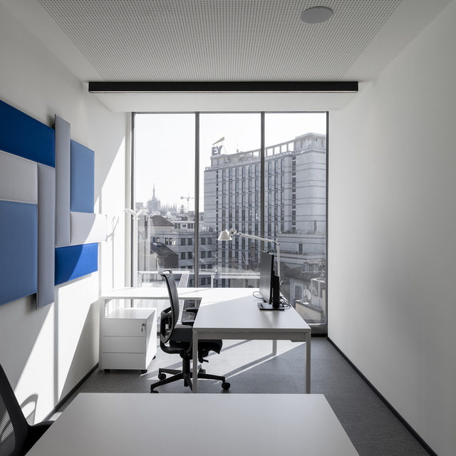 Postazione ufficio moderna con lampada da tavolo Artemide Tolomeo su scrivania bianca, sedia ergonomica, grande finestra con vista città