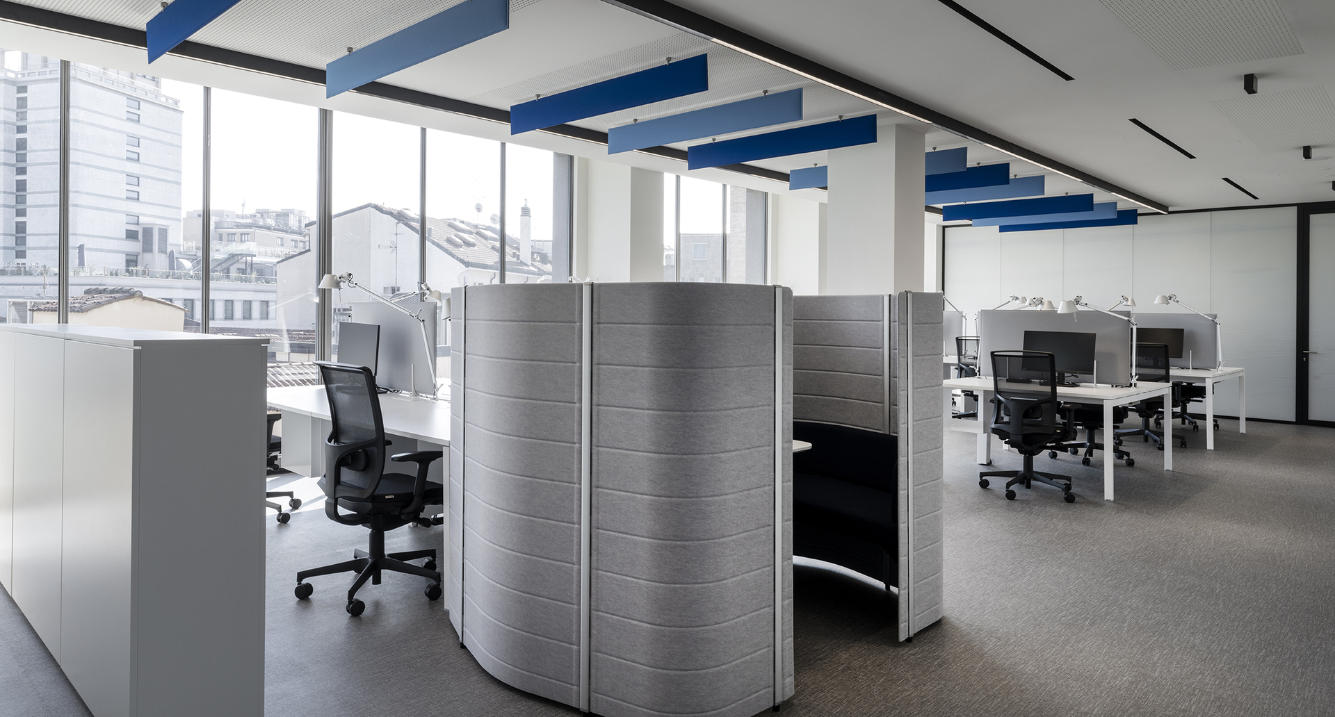 Ufficio open space moderno con postazioni di lavoro, divisori acustici a pod, sedie ergonomiche e luci lineari da incasso Artemide.