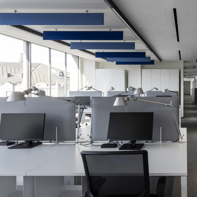 Ufficio open space moderno con lampade da tavolo Artemide Tolomeo sulle postazioni e pannelli acustici blu sospesi dal soffitto