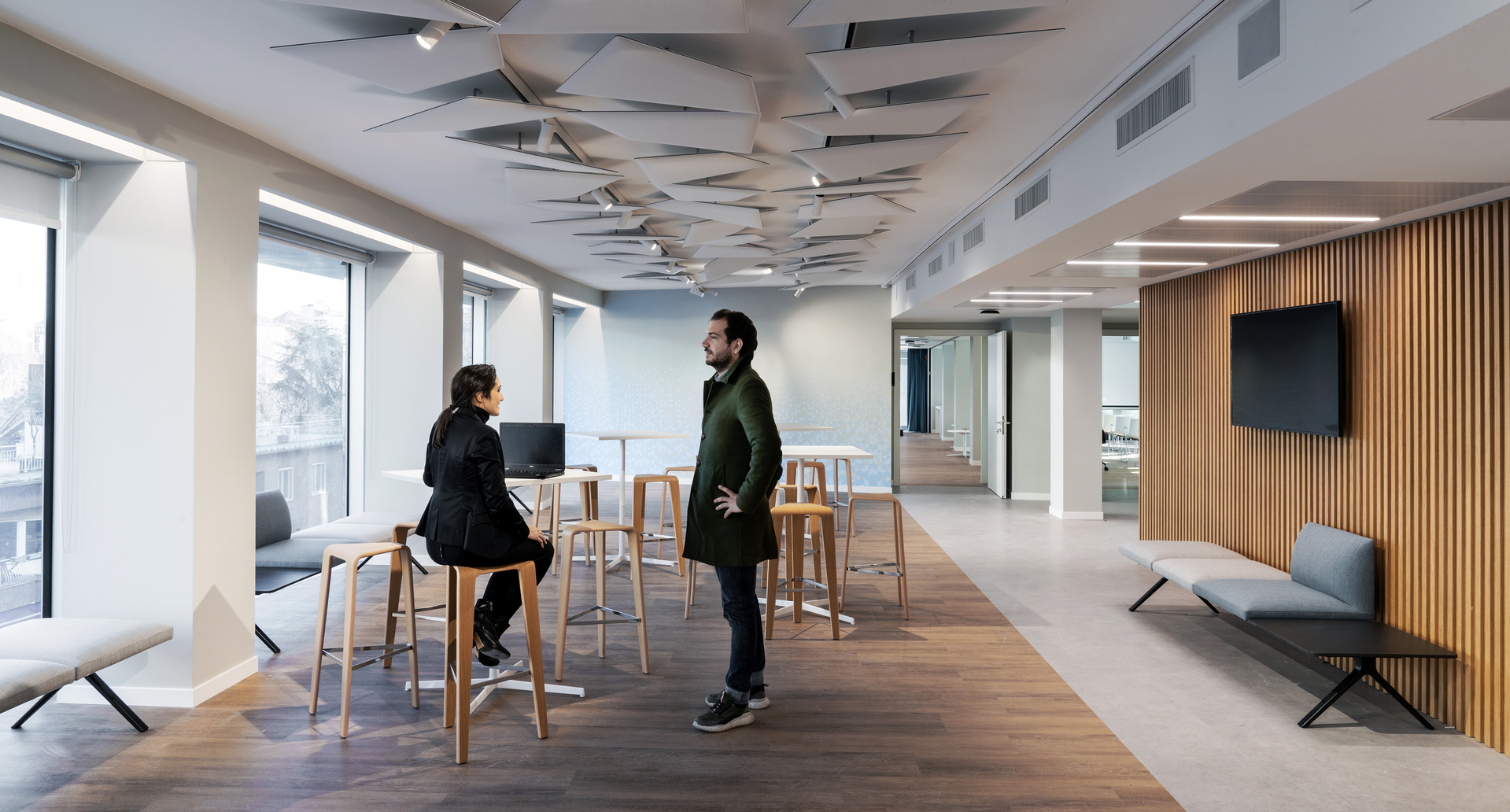 Lounge ufficio moderno con due persone in conversazione vicino a tavoli alti, con pannelli decorativi a soffitto e illuminazione LED lineare.