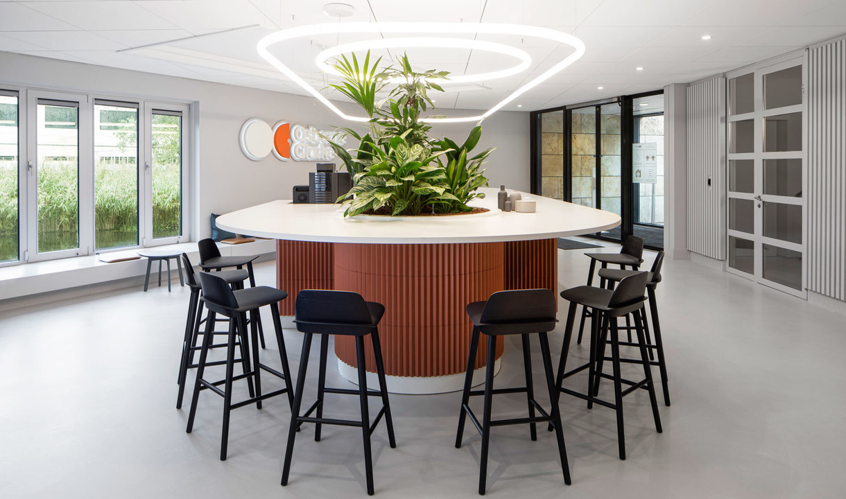 Lounge ufficio moderno con lampada a sospensione triangolare Artemide sopra un tavolo rotondo con piante e sgabelli neri.