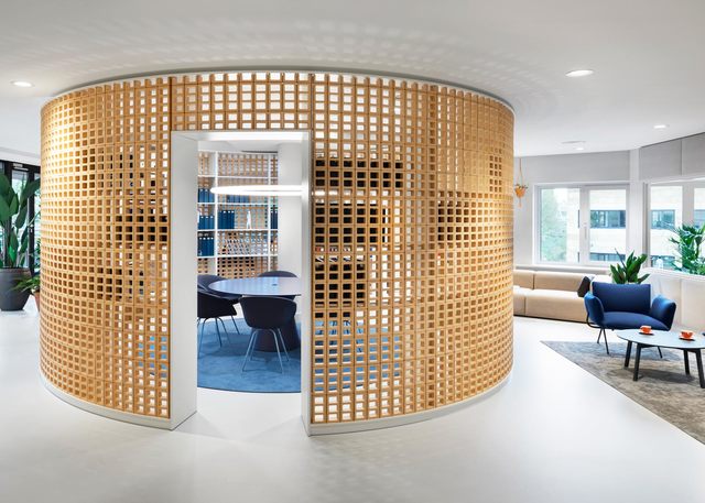 Lounge ufficio moderno con divisorio a griglia in legno curvo che racchiude una sala riunioni, sedute blu e luci a soffitto incassate.