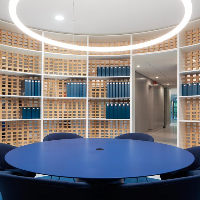 Sala riunioni circolare con tavolo rotondo blu, sedie blu navy, scaffalature a griglia in legno e lampada ad anello Artemide a sospensione.