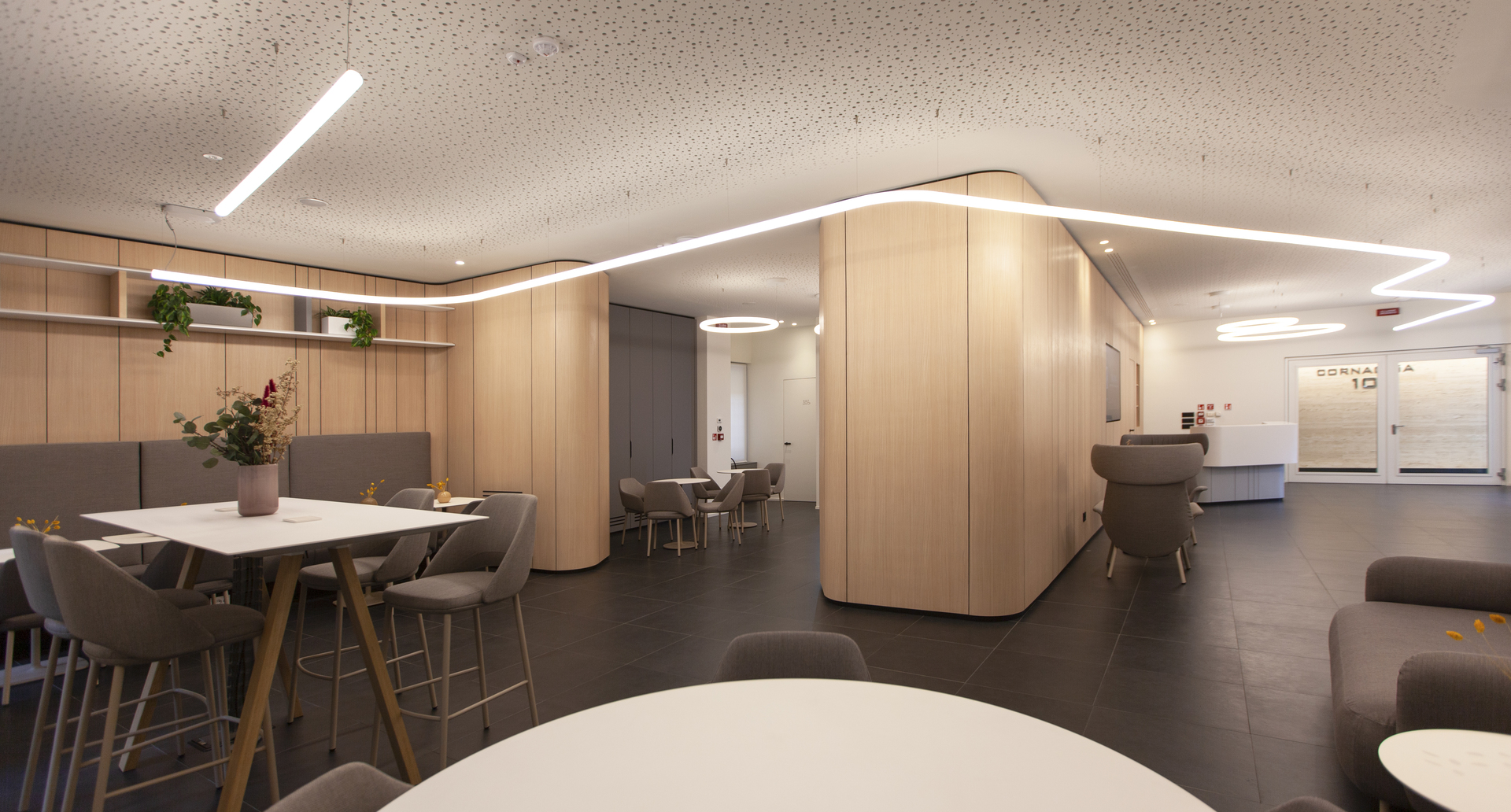 Modern office lounge with curved wood partitions, linear LED ceiling lights, pendant ring lamp, and grey upholstered seating areas