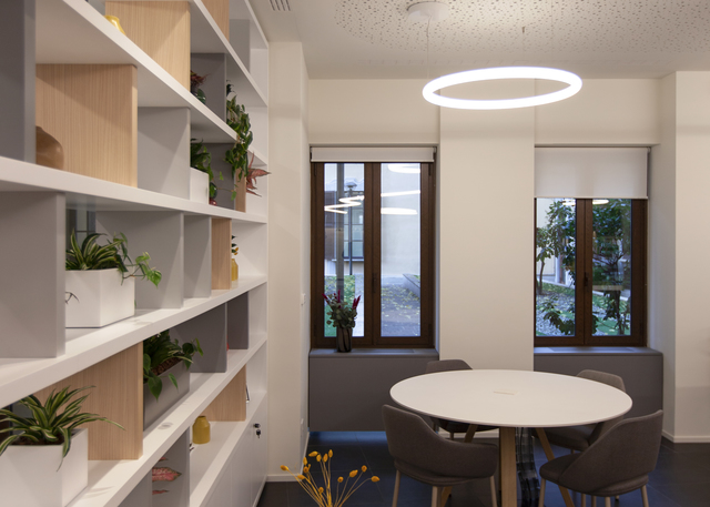 Meeting room with Artemide circular pendant light over a round white table, chairs, and a bookshelf with plants by windows.