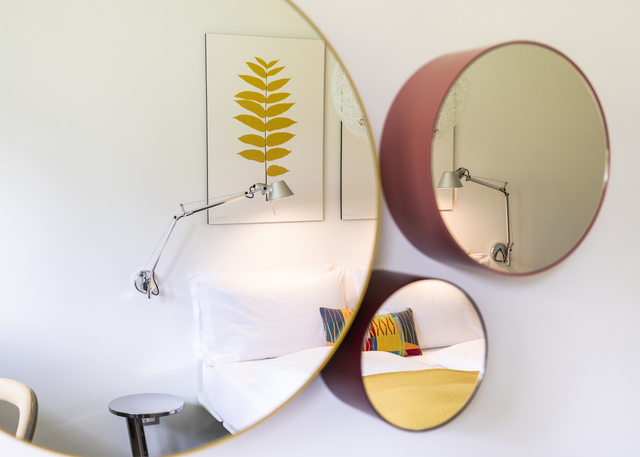 Bedroom with three round mirrors reflecting an Artemide Tolomeo wall lamp, white bedding, colorful pillows, and botanical wall art.