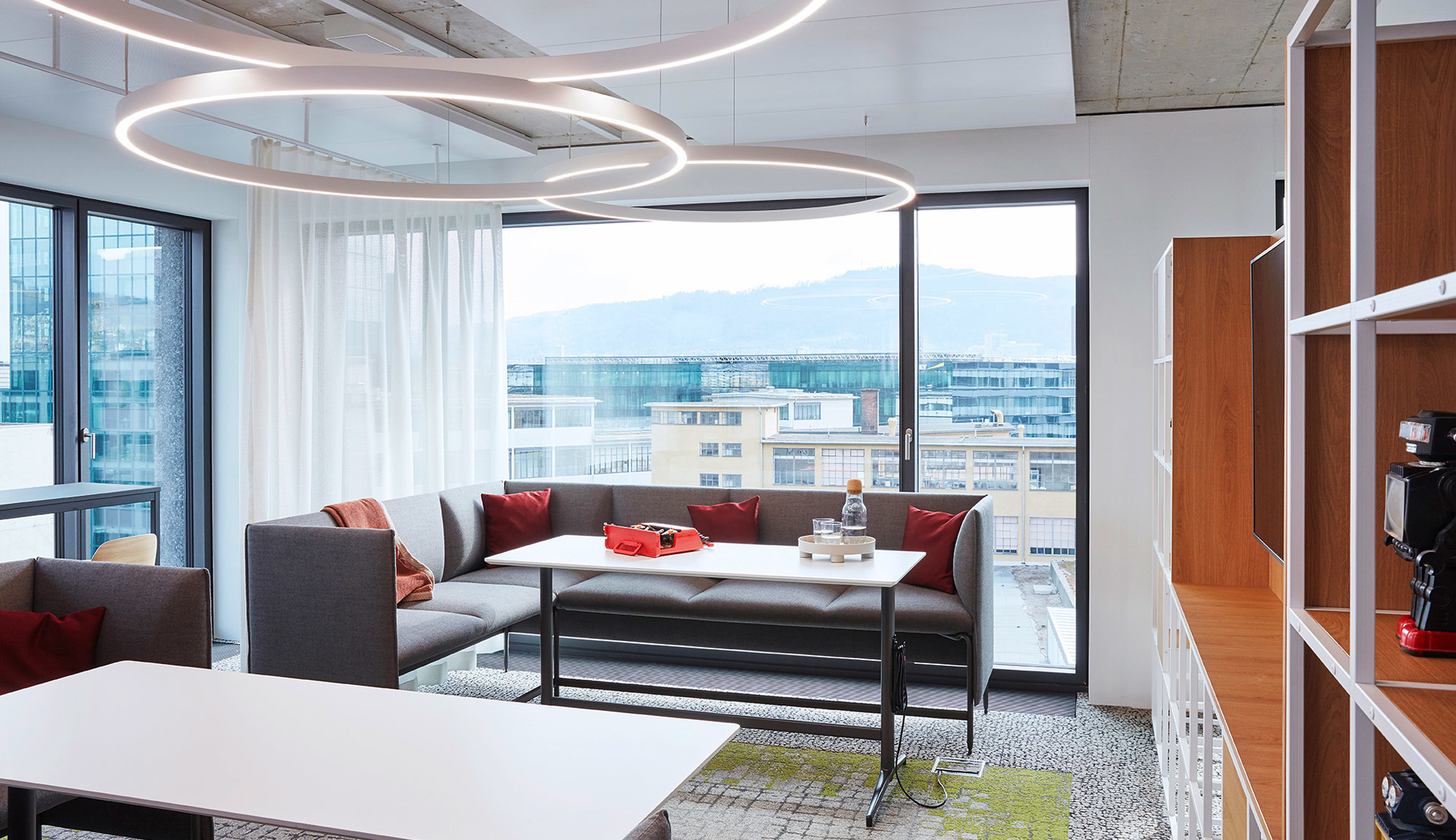 Lampada a sospensione ad anello circolare Artemide illumina un lounge ufficio moderno con divani grigi, cuscini rossi e vista panoramica sulla città.