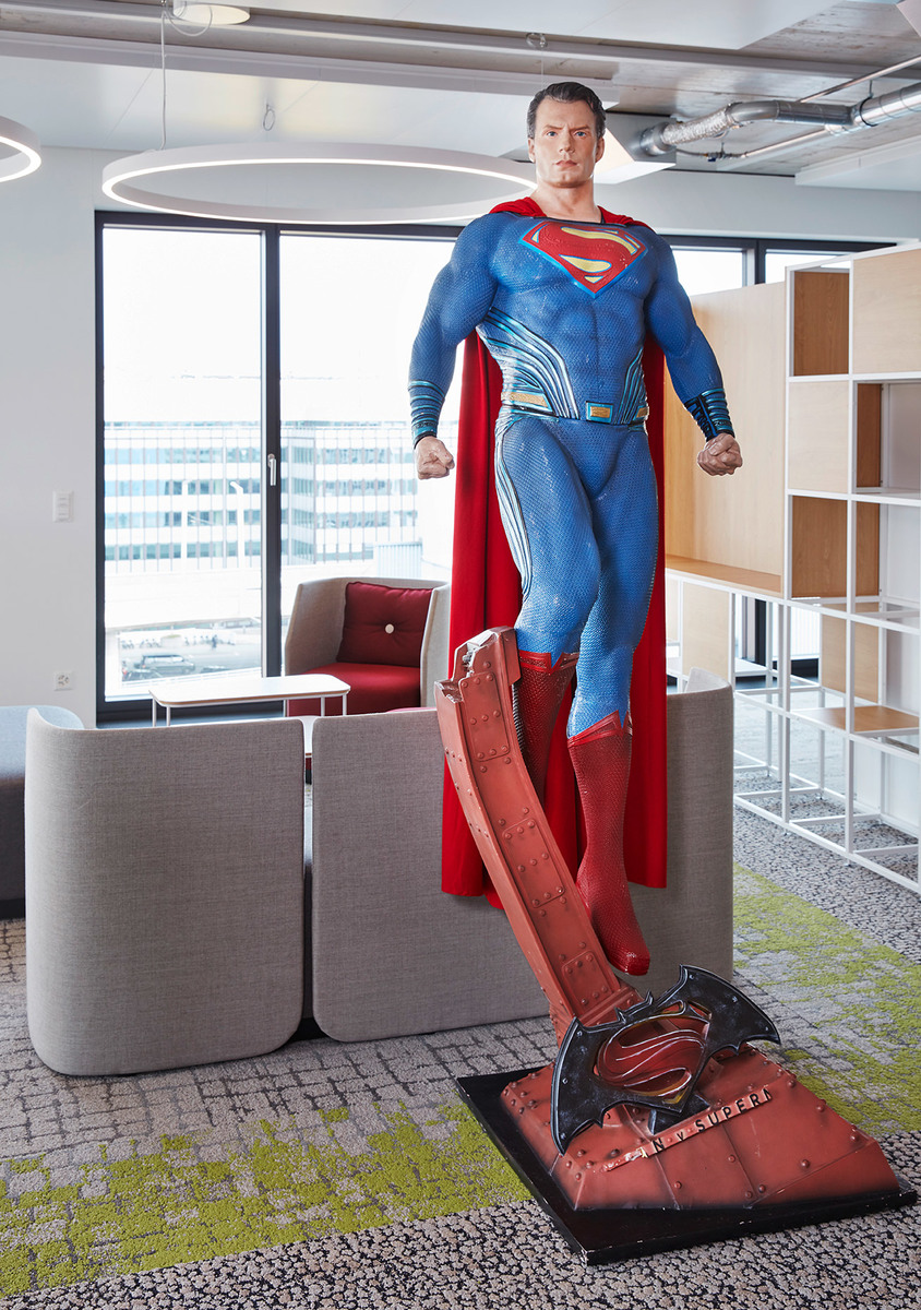 Statua a grandezza naturale di Superman in un lounge ufficio moderno con luce circolare a soffitto, divani grigi e libreria aperta.