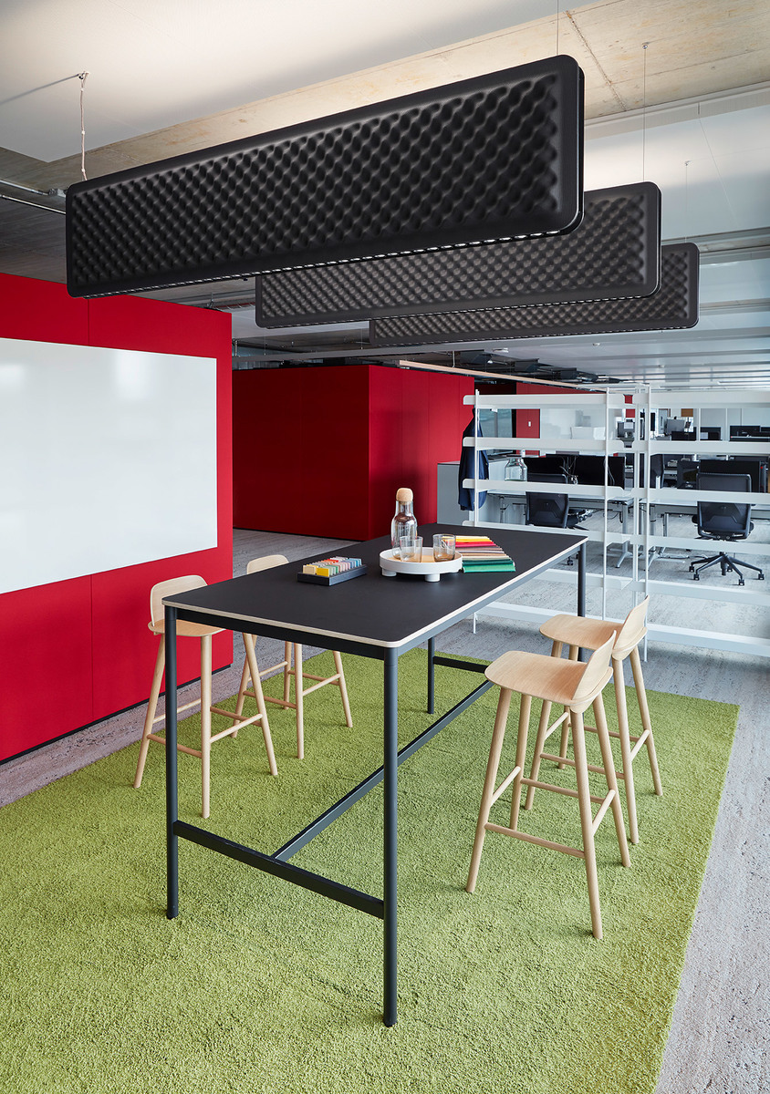 Area relax ufficio moderna con tavolo alto, sgabelli in legno, moquette verde, pareti rosse e pannelli acustici neri a soffitto.