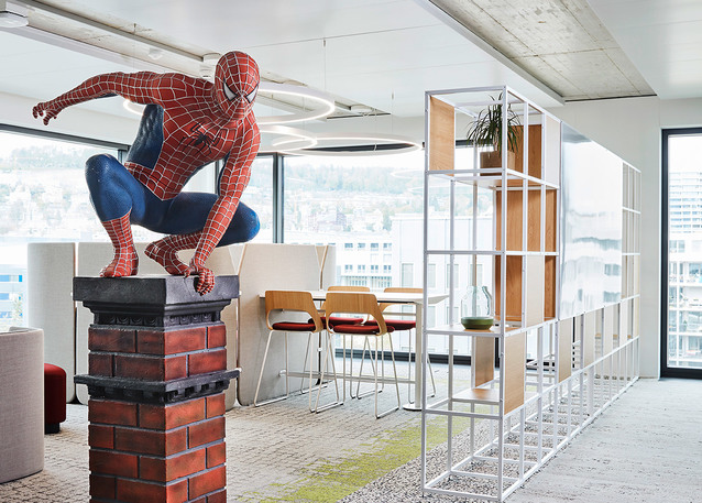 Ufficio moderno con statua di Spider-Man su piedistallo in mattoni, libreria divisoria aperta, zona pranzo e plafoniera circolare a soffitto.