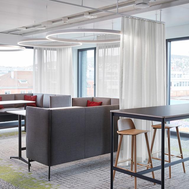 Lounge ufficio moderno con lampade a sospensione circolari Artemide sopra divani grigi, cuscini rossi e un tavolo alto con sgabelli in legno.