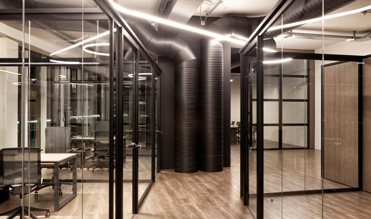 Modern office corridor with glass partitions, wood floors, dark exposed ducts, and linear LED ceiling lights illuminating meeting rooms.