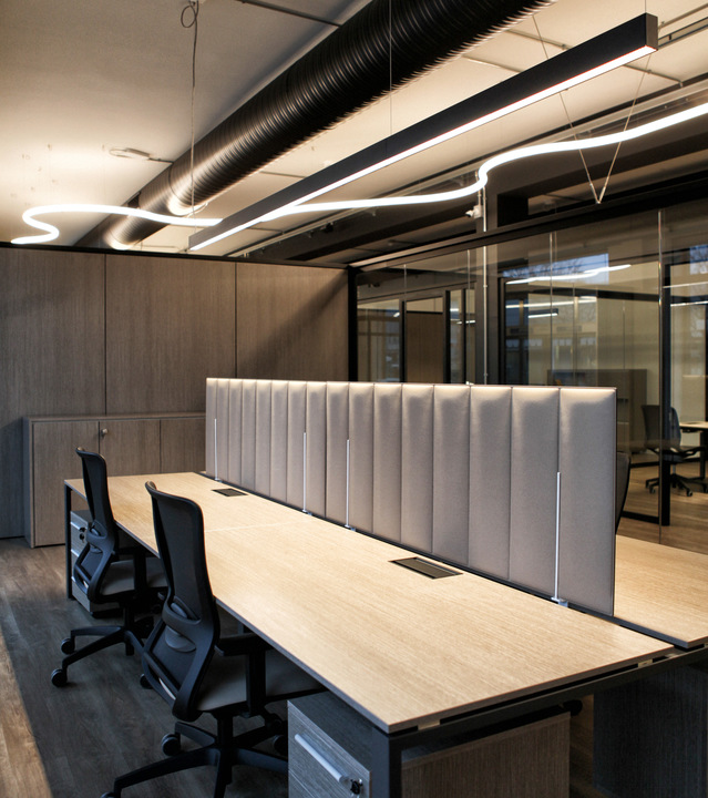 Modern office workspace with wooden desks, acoustic dividers, and sinuous white LED pendant lights suspended from an industrial ceiling.