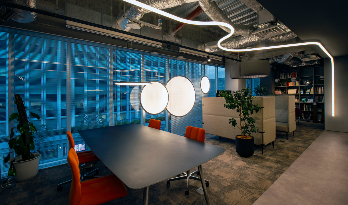 Sala riunioni ufficio moderno con lampade a sospensione circolari Artemide e striscia LED sinuosa a soffitto sopra tavolo scuro con sedie arancioni