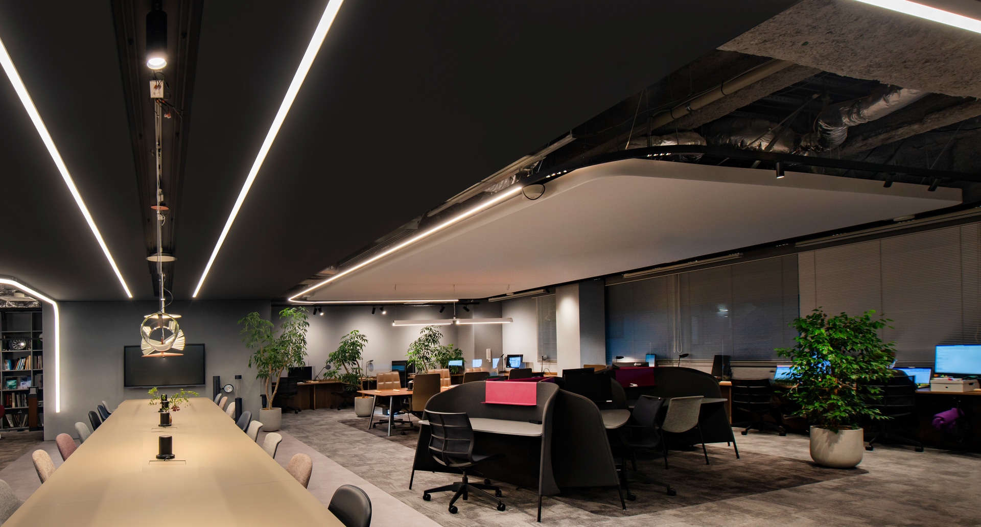 Ufficio moderno con soffitto scuro e strisce LED lineari che illuminano un lungo tavolo conferenze e postazioni di lavoro aperte con piante.