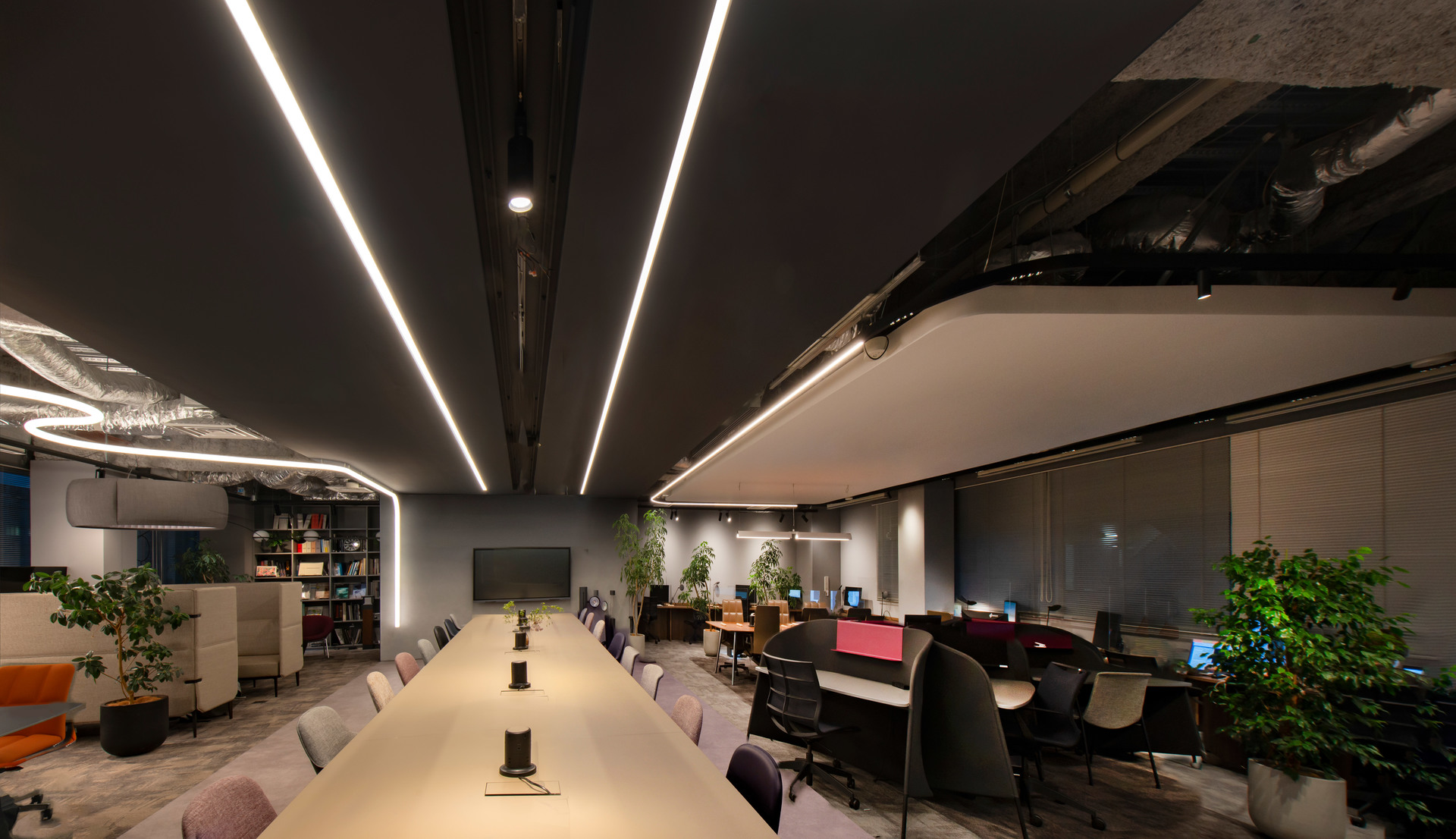 Ufficio moderno con lungo tavolo conferenze illuminato da luci lineari LED a soffitto, postazioni lavoro, piante e soffitto a vista scuro.