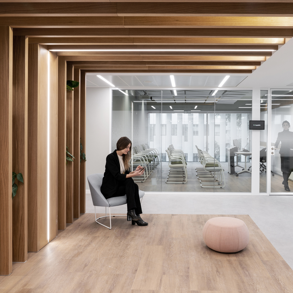 Modern office lounge with warm wood-paneled corridor, integrated LED strip lighting, and glass-partitioned workspace beyond.